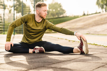 Young flexible sportsman stretching in fitness outfit on sporting ground