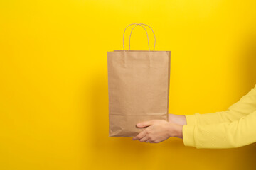 Female hands hold large gift bag made of brown craft paper on yellow background.
