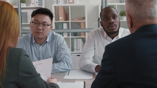 Medium close-up of two diverse job applicants sitting at table in office. African guy and Asian young man talking to unrecognizable interviewers