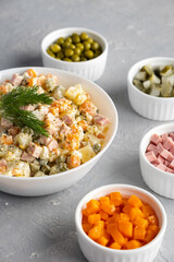 Traditional Russian Olivier salad in a white bowl and main ingredients green peas, boiled carrots, ham, pickles on a gray background. Closeup, no people