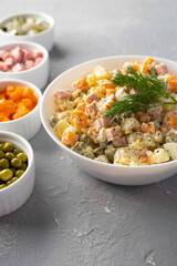 Traditional Russian Olivier salad in a white bowl and main ingredients green peas, boiled carrots, ham, pickles on a gray background. Closeup, no people