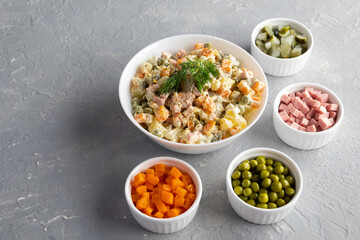 Traditional Russian Olivier salad in a white bowl and main ingredients green peas, boiled carrots, ham, pickles on a gray background. Top view, copy-space, no people