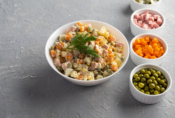 Traditional Russian Olivier salad in a white bowl and main ingredients green peas, boiled carrots, ham, pickles on a gray background. Top view, copy-space, no people