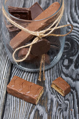 Chopped up chocolate bar. Filled with caramel and nuts. Nearby is a glass jar with chocolates. On black pine boards.