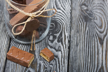 Chopped up chocolate bar. Filled with caramel and nuts. Nearby is a glass jar with chocolates. On black pine boards.