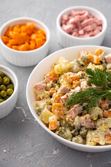 Traditional Russian Olivier salad in a white bowl and main ingredients green peas, boiled carrots, ham, pickles on a gray background. Closeup, no people