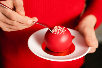 Beautiful young female hands holding red ball mousse dessert decorated with white snowflake on white plate. Christmas celebration concept. Winter season backdrop for your design.