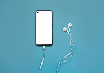 Modern phone with a white screen with headphones on a colored blue background. Concept of listening to music using a smartphone. view from above. Flat lay. copy space