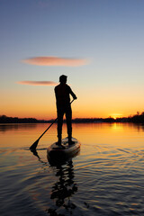 Rear view on silhouette of stand up paddle boarder paddling at sunset on river at cold time