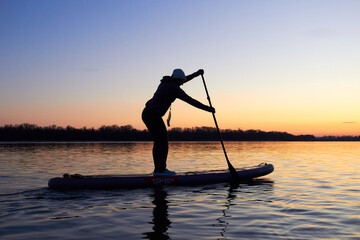 Woman on stand up paddle boarding at dusk on a flat quiet winter river with beautiful sunset colors - obrazy, fototapety, plakaty