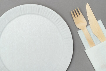 Top view of white paper dish, wooden fork and knife on white paper napkin on gray background. Set...