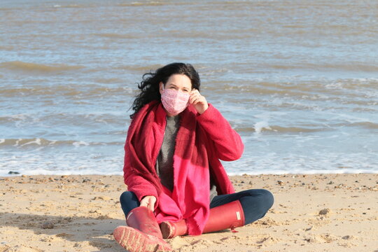 Portrait of woman in mask sat on sand on beach landscape by ocean shore wearing red with cross legs, the black haired attractive female happy in fresh air in lockdown for covid-19 in Norfolk England