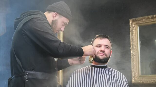 Retro styled barbershop and professional hairstylist shaving his client's head in smokey and atmospheric room with mirrors. Caucasian barber dressed in black clothing with hat shaves his client using