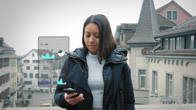 Woman stands on bridge between buildings and checks business data and graphs on her smartphone. Digital overlay display. Futuristic HUD hologram.