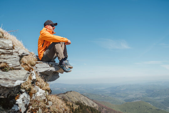 Active people and European tourism concept image. Dressed bright orange jacket hiker in baseball cap and sunglasses sitting on rocky cliff enjoying green valley at Mala Fatra mountain range,Slovakia