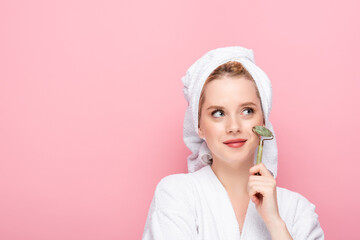 dreamy young woman in bathrobe with towel on head using jade roller isolated on pink.