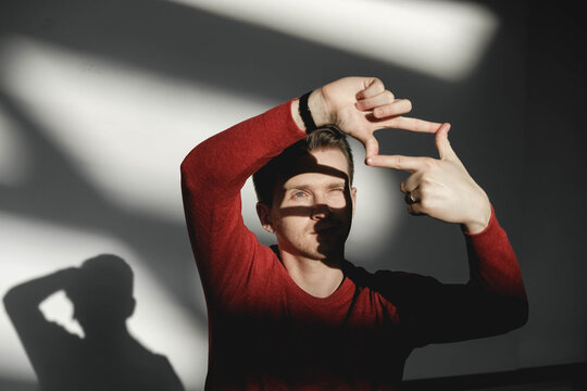 Handsome man looking through a frame formed by his hands in a red sweater on a white background with shadows from the light. Copy, empty space for text