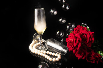 Glass of champagne, red roses, pearl necklace and soap bubbles, black background, selective focus.