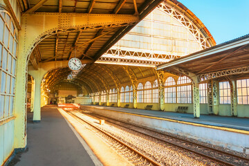 View of the old railway station with a large metal arch and an empty platform with a clock for people on a clear sunny day. © aapsky