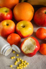 Fresh red-orange and yellow fruits lie on a gray canvas, in front of a bitten apple are open vitamins