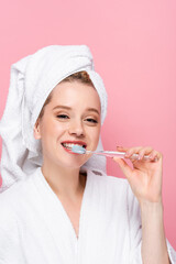 smiling young woman in bathrobe with towel on head brushing teeth isolated on pink.