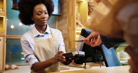 Close up of African American pretty female seller standing in bakery shop at counter typing on computer selling food. Customer hand paying with credit card on device buying baking in bakehouse