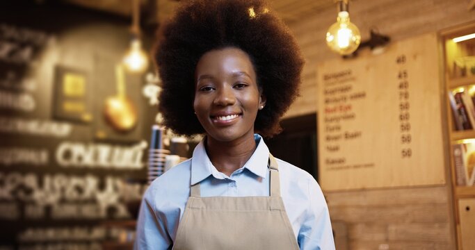 Close up portrait of happy beautiful African American young woman entrepreneur in apron standing in own small bakery shop, looking at camera and smiling in bakehouse. Business concept