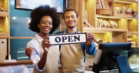 Portrait of cheerful young African American family married couple of bakers in aprons reopening bakehouse holding Open sign in hands. Small own bakery shop. Business concept - Powered by Adobe