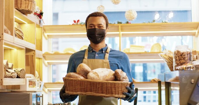 Close up of handsome happy young African American man bakery worker in face mask standing in bakehouse and holding basket with fresh baked bread. Bakery owner looking at camera and smiling