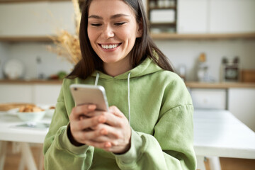 Happy positive student girl in green hoodie holding cell phone, smiling broadly, flirting with her...