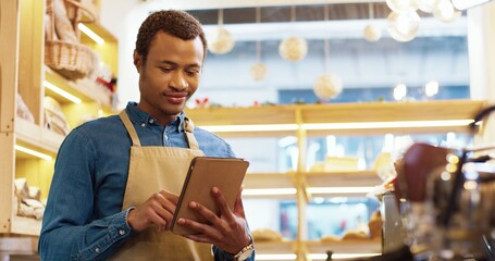 Close up of African American happy young handsome man worker in apron stands in bakery shop, texting and typing on tablet. Male seller browsing online on device. Business concept. Retail industry - obrazy, fototapety, plakaty