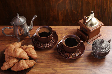 Two cups of coffee, coffee pot, coffee grinder, croissants on a saucer, saucer with cookies and chocolates on wooden texture.