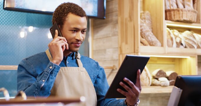 Close up of joyful African American young handsome man worker in apron stands in bakery shop, speking on smartphone and using tablet device. Business concept. Retail industry