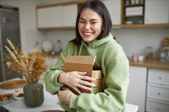 Cheerful teenage girl expressing excitement being overjoyed after receiving long awaited parcel with new clothing or accessories, embracing cardboard box, smiling broadly with her eyes closed