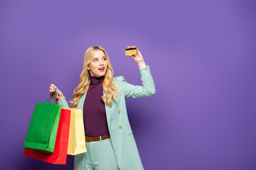 blonde young woman in fashionable turquoise blazer with shopping bags and credit card on purple background.