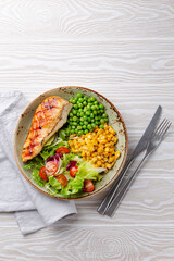 Baked chicken breast on plate with fresh salad, green peas and corn, white wooden background, top view. Healthy fitness meal with chicken fillet, balanced in proteins and carbs. Overhead, copy space