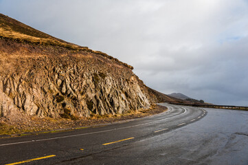 Dark wet road near sun lit cliffs going from left to right in Ireland near Atlantic ocean in a rainy day in winter and autumn. Concepts: season, outdoors, travelling, landscape