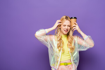 stressed blonde young woman in colorful outfit with paper cup on purple background.