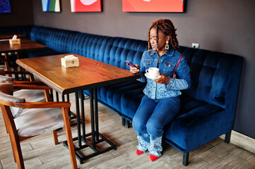 Lovely african american woman with dreadlocks in blue stylish jeans jacket at cafe hold mobile phone. Beautiful cool fashionable black young girl indoor drink coffee.