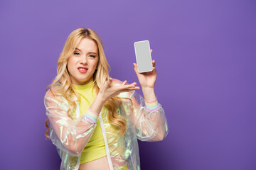 confused blonde young woman in colorful outfit presenting smartphone on purple background.