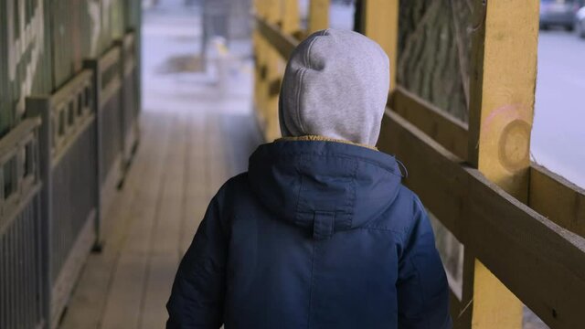 A homeless child waif walks along a fence in a slum. The view from the back. An orphan boy walks down the street