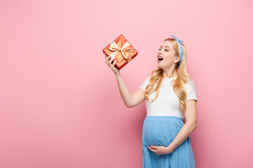 happy blonde young pregnant woman with gift box on pink background.