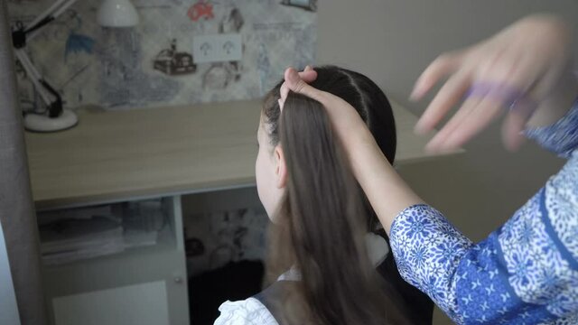 Mom combs her hair and makes ponytails on the head of a little schoolgirl girl who is sitting in front of her on a chair in the room. The concept of mother's care. Close-up. Rear view. 4K