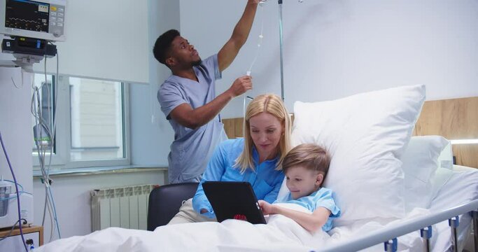 Cheerful Caucasian caring mother sitting with little happy boy kid tapping on tablet device screen in hospital room and smiling while African American male nurse putting a dropper. Treatment concept