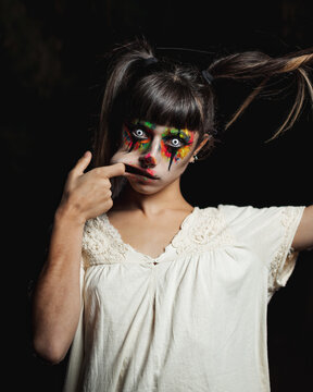 Scary young female in colorful contact lenses and with painted face standing in dark forest and smiling creepy while looking at camera on Halloween