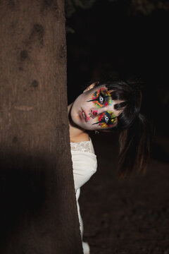 Creepy female with creative makeup and in contact lenses standing near tree in woods at night on Halloween and looking at camera