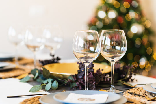 Dining table served with glasses and plates against blurred glowing Christmas tree on Christmas eve at home