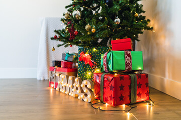 Heap of colorful ornamental present boxes with ribbons near decorative fur tree and shiny letters on floor with garland during festive event