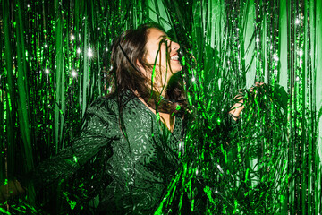 Delighted female wearing festive dress playing with green shiny tinsel stripes at party while looking at camera