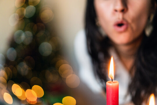 Crop anonymous female blowing on shiny wax candle fire near decorative Christmas tree at home